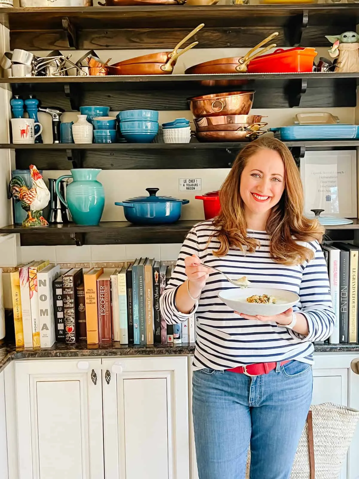 Anina Giannini in her kitchen, smiling while holding a bowl in front of shelves of French cookware and cookbooks